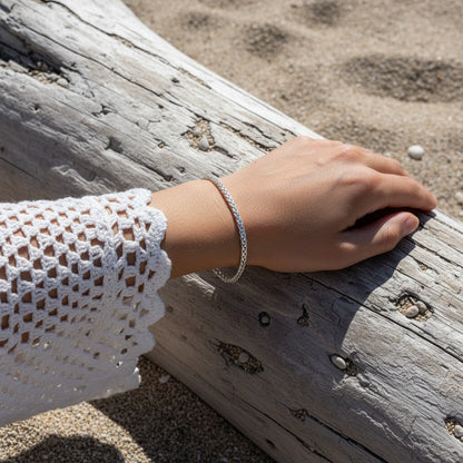 Aramat Jewels zilverkleurige vossenstaart armband van RVS gedragen door een persoon op een strand bij een boomstam.